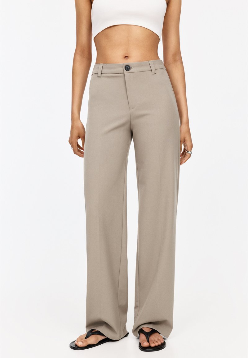 Pantalon large beige au toucher lisse, avec une fermeture à bouton, des passants de ceinture et une coupe ajustée. Associé à des tongs noires.