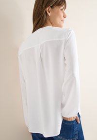 Blouse blanche avec une texture douce et transparente, manches longues, ourlet arrondi, et un design simple. Le dos présente un détail de panneau lisse.