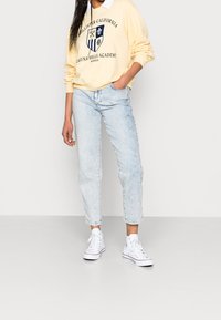 Person som bär en ljusgul sweatshirt med en sköldlogotyp, ljusblå jeans och vita högstövlar, står mot en plain ljusvägg.