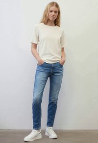 Young person standing with hands in pockets wearing a cream short-sleeve shirt, blue jeans, and white sneakers against a plain wall.