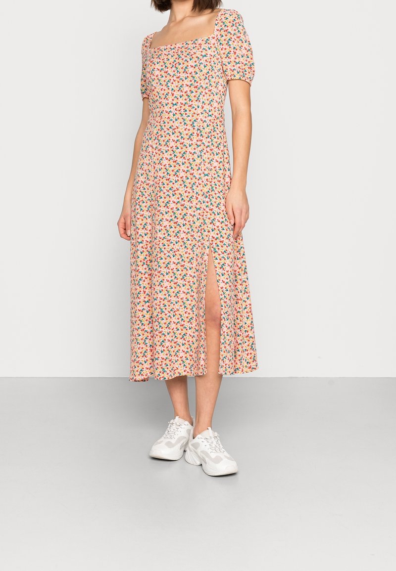Femme portant une robe midi à fleurs avec des manches bouffantes et une fente frontale, associée à des baskets blanches épaisses, debout devant un fond uni.