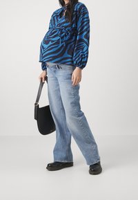 Blouse à rayures zébrées bleu et noir avec manches bouffantes, associée à un jean évasé bleu clair et des sabots noirs ; tenant un sac à bandoulière noir.
