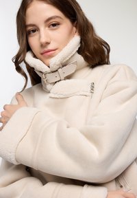 Cappotto in pile beige con colletto alto, dotato di un interno foderato in pelliccia, chiusura con cerniera e maniche ampie. Rifinito con un dettaglio a fibbia in vita.