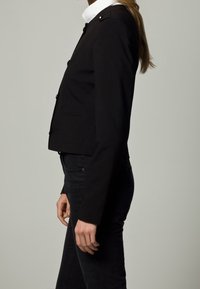 Černá zkrácená bunda s elegantním designem, zaobleným límcem, předními kapsami a knoflíkovými detaily na ramenou.