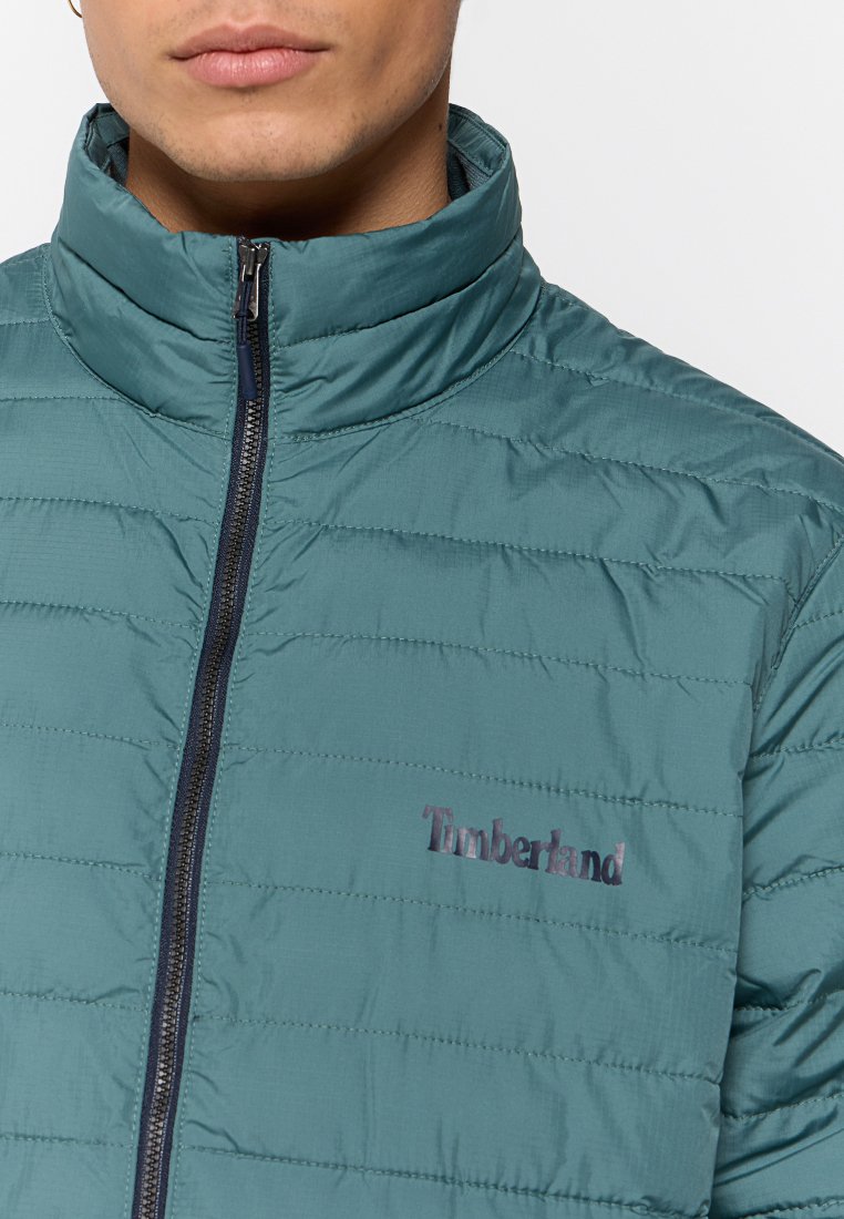 Türkisfarbene, gepolsterte Jacke mit hohem Kragen, Reißverschluss und gesteppter Textur. Verfügt über ein kleines, gesticktes Timberland-Logo auf der Brust.