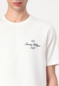 T-shirt en coton blanc avec des manches courtes, un col rond et un petit logo noir avec "Tommy Hilfiger" et "N.Y.C." imprimé sur le devant.