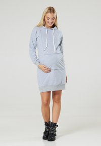 Hellgraues Umstandshoodiekleid mit langen Ärmeln, einer Kängurutasche und Zugbändern. Das Kleid hat einen gerippten Saum und wird mit kontrastierenden schwarzen, genagelten Stiefeln kombiniert.
