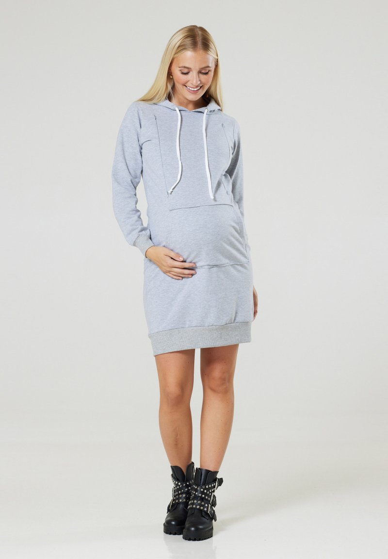 Hellgraues Umstandshoodiekleid mit langen Ärmeln, einer Kängurutasche und Zugbändern. Das Kleid hat einen gerippten Saum und wird mit kontrastierenden schwarzen, genagelten Stiefeln kombiniert.
