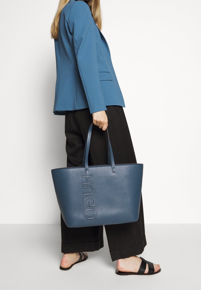 Personne portant un blazer bleu et un pantalon noir tenant un grand sac cabas bleu avec "HUGO" embossé dessus, portant des sandales noires.
