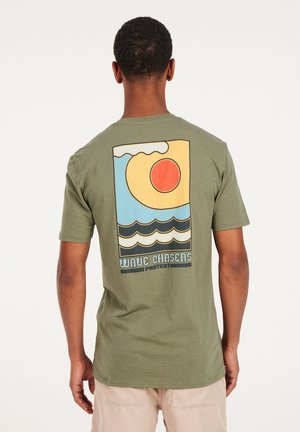 Olijfgroene katoenen t-shirt met een grafisch ontwerp op de achterkant, met een zon, golven en de tekst "WAVE CHASERS" in dikke, contrasterende kleuren.