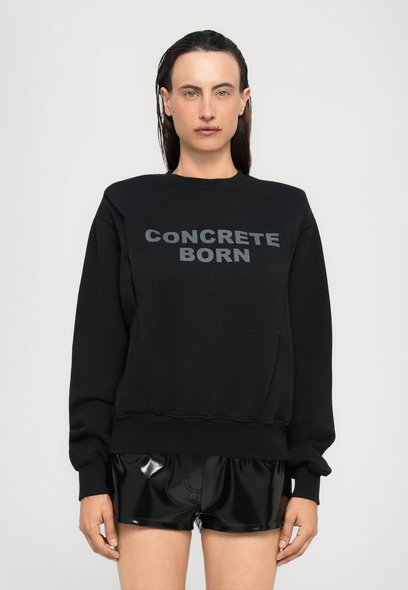 Persona con capelli bagnati che indossa una felpa nera con la scritta "CONCRETE BORN" e pantaloni corti neri lucidi, in piedi contro uno sfondo chiaro e uniforme.