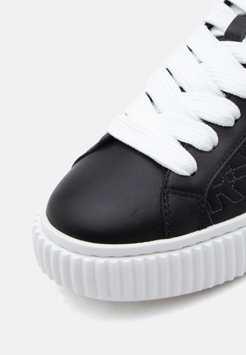 Zapatilla de cuero negra con suela de goma blanca, con estrías gruesas, cordones blancos planos y un patrón sutil en relieve en la parte superior.