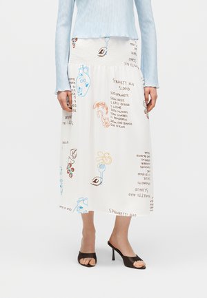 Jupe midi blanche avec illustrations alimentaires colorées dessinées à la main et texte de recette, portée avec un haut bleu clair et des sandales à talons noires.