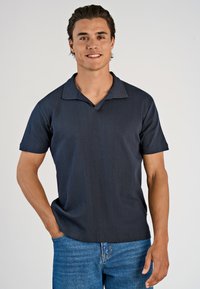 Hymyilevä tummahiuksinen mies, jolla on yllään tummansininen lyhythihainen polo-paita ja siniset farkut, seisoo yksi käsi taskussa tasaisella taustalla.