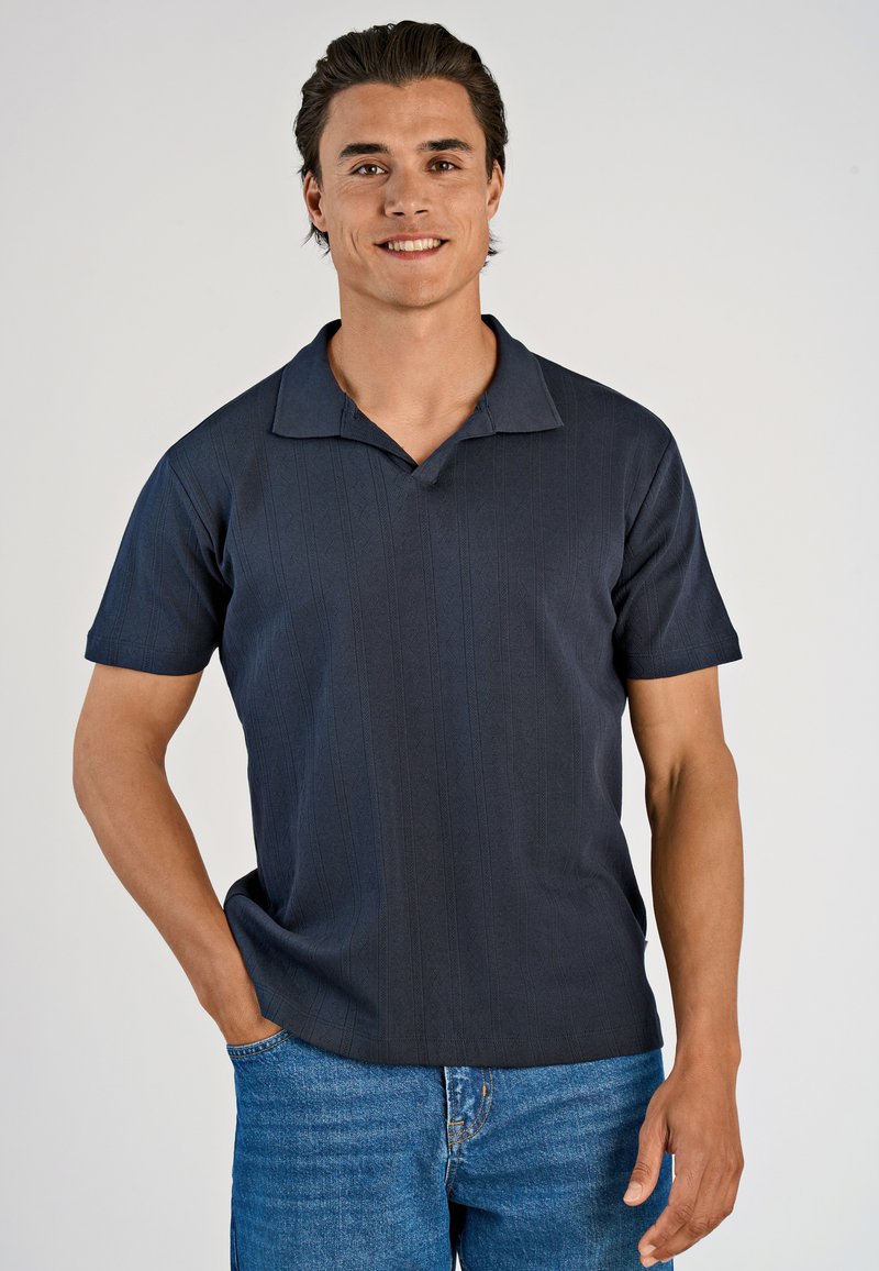 Hymyilevä tummahiuksinen mies, jolla on yllään tummansininen lyhythihainen polo-paita ja siniset farkut, seisoo yksi käsi taskussa tasaisella taustalla.