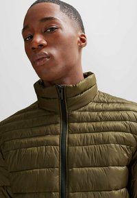 Chaqueta tipo puff en verde oliva con cuello alto, cierre de cremallera, textura acolchada y un acabado ligeramente brillante.