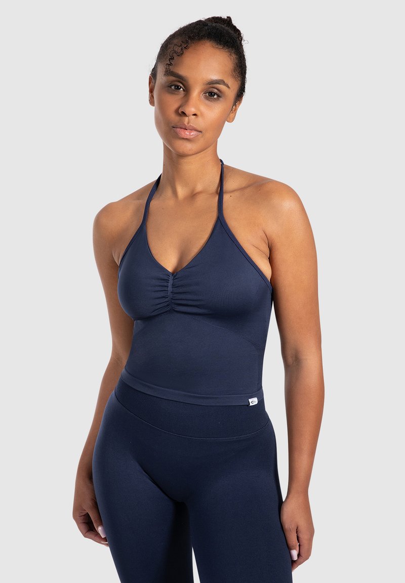 Vrouw met krullend haar, gekleed in een marineblauwe haltertop en bijpassende hoogmatige leggings tegen een effen lichtgrijze achtergrond.