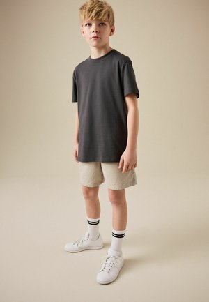 Blonder Junge steht, trägt ein dunkelgraues T-Shirt, beige Shorts, weiße Turnschuhe und weiße Socken mit zwei schwarzen Streifen, neutraler Hintergrund.