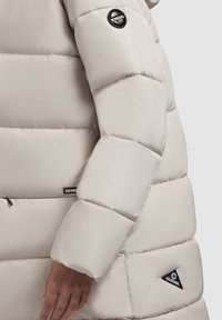 Pufferjack in lichtbeige, met een gevoerd ontwerp en een glanzende stoftextuur. Inclusief zwarte logopatches op de mouw en zijkant.