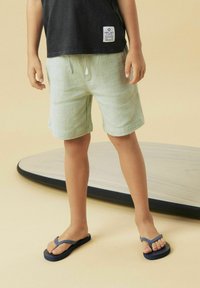 Shorts texturés vert clair avec une taille élastique et un cordon de serrage, associés à des tongs bleu foncé. Le fond présente une planche courbée.