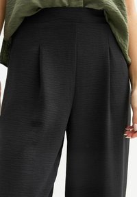 Zwarte plooibroek van een gestructureerde stof, met een hoge taille en een comfortabele pasvorm. Gepaard met een losse groene top.