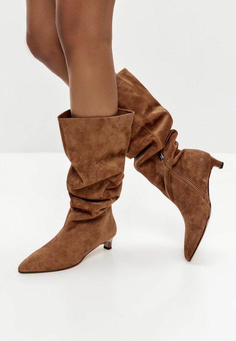 Bruine suede slouch boots met een spitse neus, middelhoge blokhak en zichtbare ritsdetails aan de zijkant. Geverfde en zachte afwerking.