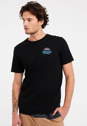 Jonge man met middellang krullend bruin haar, draagt een zwart T-shirt met een regenbooglogo en de tekst 'Protest' op de borst, kijkt naar links.