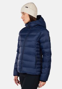 Marineblå puffjakke med hætte, med quiltede sektioner, lynlåslommer og en glat, blank overflade. Parret med en cremefarvet strikbeanie.