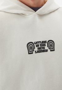 Sudadera con capucha en tejido gris claro que presenta un estampado gráfico en negro con el texto "ESTAR FUERA DE LÍNEA ES EL NUEVO LUJO" y diseños de cabezas abstractas.