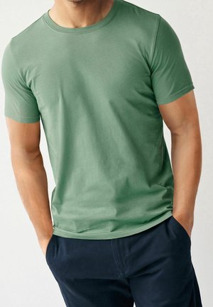 Homme portant un t-shirt à manches courtes vert clair uni et un pantalon bleu marine foncé, les mains dans les poches, se tenant devant un fond clair.