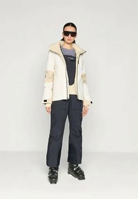 Γυναίκα που φοράει γυαλιά ηλίου, μπεζ και άσπρο χειμερινό μπουφάν, μαύρες παντελόνες χιονιού και σκιέρ μπότες, στέκοντας μπροστά σε ένα απλό άσπρο φόντο.