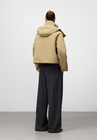 Osoba z włosami w kok w beżowej kurtce z kapturem i szerokich ciemnoszarych spodniach, stojąca twarzą do białej ściany, z rękami w kieszeniach.