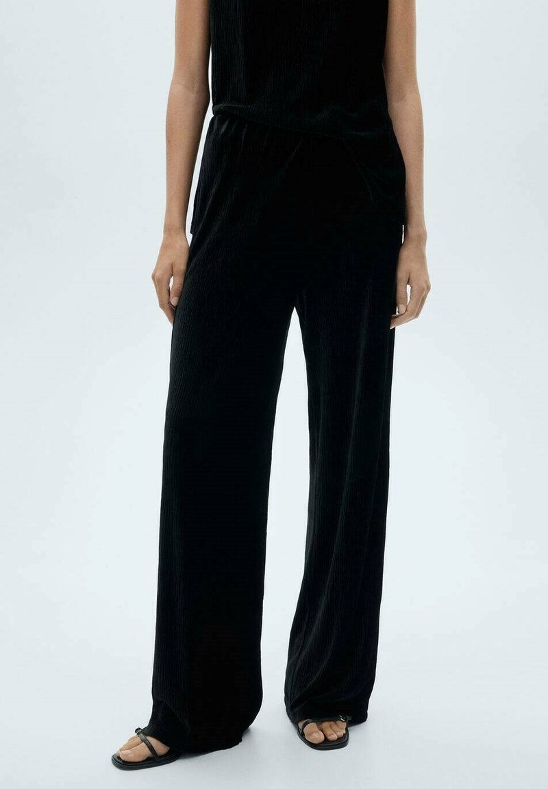Pantaloni negri cu croială largă, din material ribbed, cu talie elastică, asortați cu sandale negre cu barete. Textură netedă cu un design fluid.