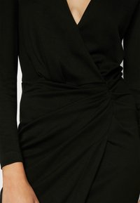 Robe portefeuille noire à manches longues avec décolleté en V et détail froncé à la taille, portée par une personne dont un bras est visible.