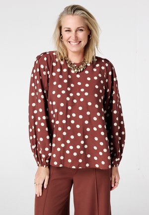 Femme souriante portant un haut à manches longues marron à pois blancs, un pantalon marron, un collier en or et des boucles d'oreilles créoles sur un fond uni.