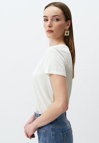 Biała koszulka z krótkim rękawem i okrągłym dekoltem, połączona z niebieskimi spodniami o wysokim stanie. Modelka nosi duże złote kolczyki.