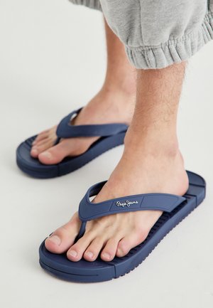 Person, der blaue Flip-Flops von Pepe Jeans und hellgraue Shorts mit elastischem Bündchen trägt, steht auf einem hellen Boden.