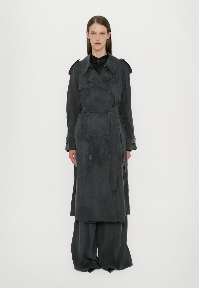 Grijze trenchcoat van suède materiaal, met dubbele knopen, een tailleband en brede mouwen, gecombineerd met bijpassende brede broek.