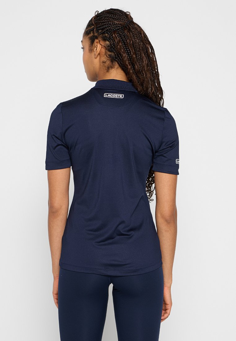 Granatowa koszulka polo, krótki rękaw, gładka powierzchnia, z logo Lacoste na tylnej części szyi i rękawie. Dopasowany krój.