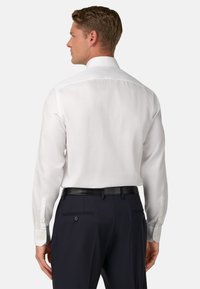 Bílá košile s dlouhým rukávem a klasickým límcem, hladká textura a knoflíkové manžety, v kombinaci s tmavými kalhotami a černým opaskem.