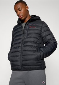 Juoda pūkuota striukė su užtrauktuku, horizontaliomis siūlėmis ir gobtuvu. Kairėje krūtinės pusėje yra raudonas "Champion" logotipas.