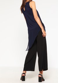 Navy blue sleeveless top with a sheer back panel, paired with loose black trousers and chunky black heeled sandals.