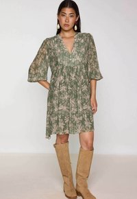 Vrouw in een groene bloemenjurk tot de knie met 3/4 mouwen en tan suède laarzen tot de knie, staand met één hand op de heup.