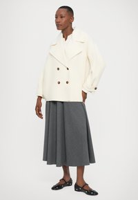 Nő off-white színű, kétsoros gombolású kabátban, szürke midi redőkben álló szoknyában és fekete Mary Jane balerinában ezüst szegecsekkel, fehér háttér előtt.