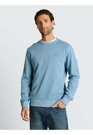 Mężczyzna z krótkimi kręconymi włosami, ubrany w jasnoniebieską bluzę i dżinsy, stojący z jedną ręką w kieszeni na jednolitym tle.