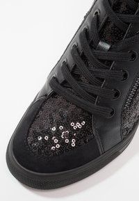Gros plan sur une basket noire avec un tissu à sequins sur l'avant, des lacets noirs, des panneaux en cuir et un détail de fermeture éclair latérale.