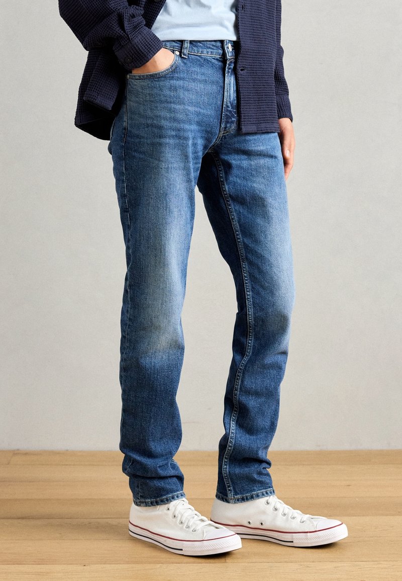 Vaqueros azules de mezclilla con acabado descolorido, diseño de cinco bolsillos y ajuste slim; combinados con zapatillas de lona blancas con detalles en rojo.