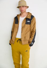 Braune und schwarze Windbreaker-Jacke mit Logo, kombiniert mit gelben Sweatpants mit Logo, die eine lockere Passform und einen Kordelzug-Bund zeigen.