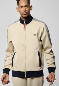 Veste de survêtement zippée beige avec un col et des poignets bleu marine, comprenant des panneaux latéraux à motifs et des poches avant. Logo sombre sur la poitrine.