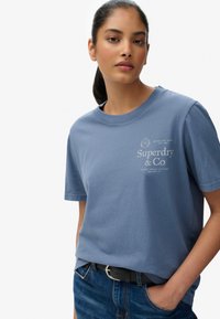 Blå bomuld T-shirt med rund hals, korte ærmer og et frontlogo til Superdry & Co. Kombineret med denimshorts og bælte.